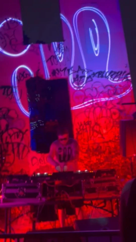 A DJ plays in front of a graffiti-covered wall under red and purple light
